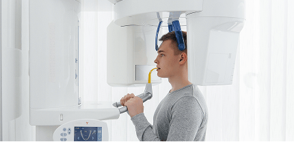 Patient undergoing a dental panoramic X-ray, standing still with a bite block in his mouth and holding support handles while the imaging machine surrounds his head.