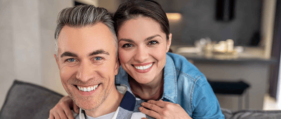 Smiling couple showing off their bright, healthy teeth, with the woman standing behind the man and resting her hands on his shoulders. Both appear happy and confident, highlighting excellent dental care.