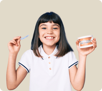 A young girl holding up a clear aligner in her right hand and a braces tooth model in her other hand.
