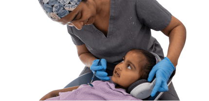 Dr. Diluni smiling and gently examining a young child’s teeth while the child wears headphones for comfort during the dental visit.