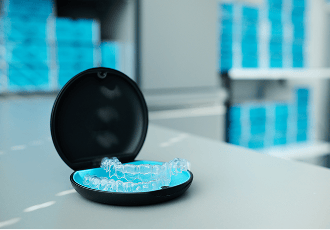 A set of clear dental aligners placed in an open black and blue protective case, resting on a table with shelves of blue clear aligner boxes in the background.