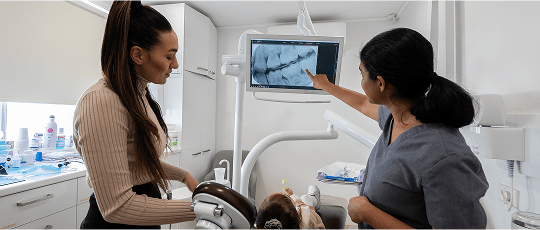 Dr. Diluni pointing to a dental X-ray on a monitor while explaining the image to a young patient and their parent in a modern clinic.