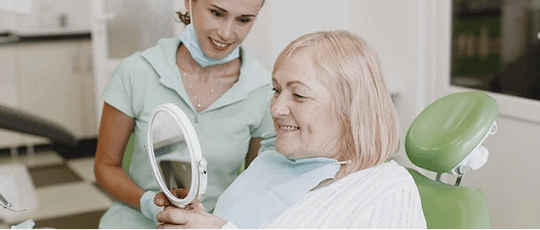 Older woman sitting in a dentist's chair, smiling as she admires her new dentures in a handheld mirror.