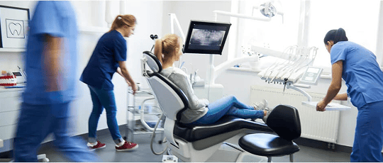 Team of dental professionals preparing for treatment while a patient sits in the dental chair, surrounded by equipment in a clinical setting.