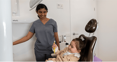 Young child holding a toothbrush and smiling while sitting in a dental chair, with Dr. Diluni nearby creating a relaxed and friendly environment.
