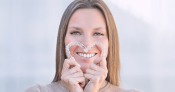 A smiling woman holding two clear aligners to form a heart shape.