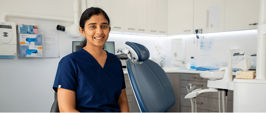 Principal dentist at Mulgrave Dental Group, Dr. Diluni Perera is smiling, seated in a modern dental clinic beside a dental chair and equipment.
