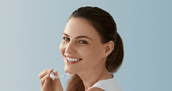 A smiling woman holds a clear aligner between her fingers, about to place it in her mouth.