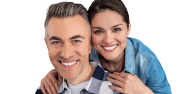 Smiling couple showing off their bright, healthy teeth, with the woman standing behind the man and resting her hands on his shoulders. Both appear happy and confident, highlighting excellent dental care.